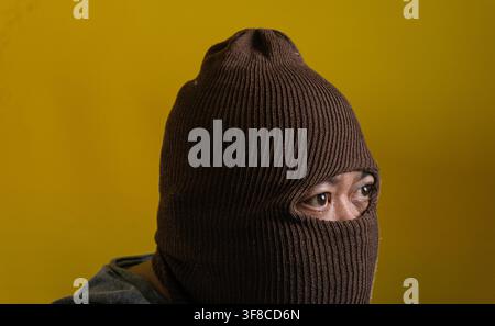 Intriguing portrait of an adult Asian male wearing a brown knitted balaclava against a bright yellow background. The anonymous individual turns slight Stock Photo