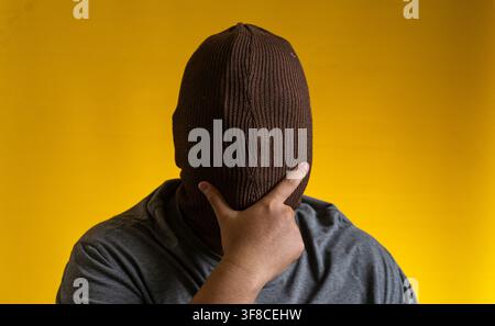 Dramatic portrait of an adult Asian male completely hiding the face with a brown knitted balaclava against a solid yellow background, resting one hand Stock Photo