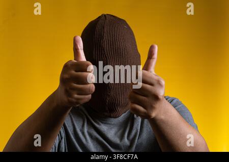 A Southeast Asian male wearing a brown knitted balaclava and a basic grey shirt gives a double thumbs up gesture directly toward the camera against a Stock Photo