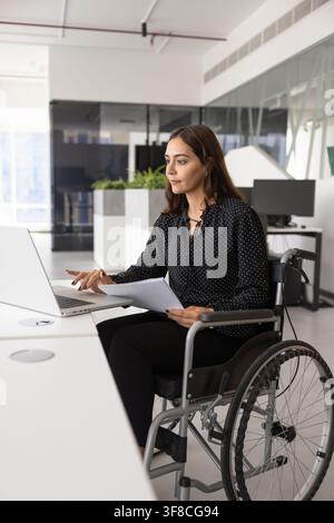 Confident woman in wheelchair using laptop at office desk Stock Photo