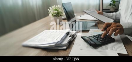 Person calculates figures on financial documents laptop while another reviews a clipboard, supporting account analysis, financial checking, business a Stock Photo
