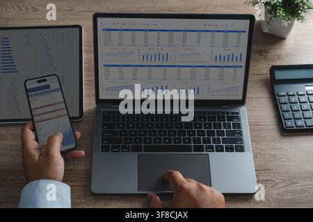 Hands use a laptop and smartphone beside a tablet and calculator, reviewing spreadsheet dashboards with charts for financial data analysis, revenue tr Stock Photo