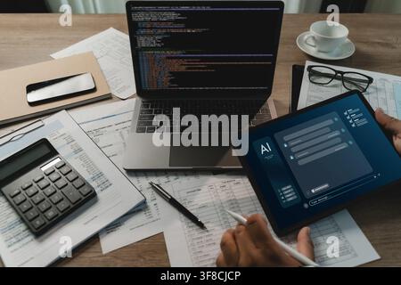 Businessman using a tablet with an AI chatbot interface beside a laptop with code, calculator and financial papers on a busy desk for digital document Stock Photo