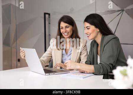 Two smiling female colleagues collaborating on digital project by laptop Stock Photo