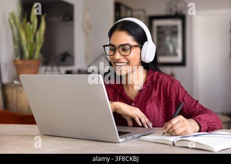 Smiling Indian student woman wearing wireless headphones studying online sits at table at home using laptop and notebook. E-learning process, remote e Stock Photo