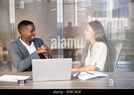 Two diverse businesspeople laughing during informal business conversation or work discussion sitting at office desk. Professional communication during Stock Photo