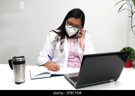Latina doctor woman works in her office very busy uses her cell phone to answer messages, calls and video calls from patients Stock Photo