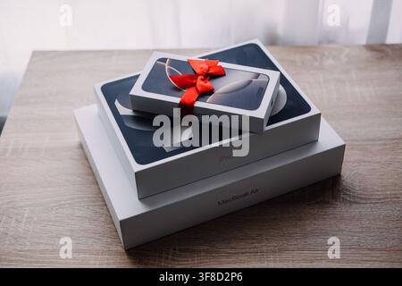 iPhone box with red ribbon bow sits on iPad Air and MacBook Air boxes on table. Apple devices with gift bow, iphone ipad macbook gift set, tech bundle Stock Photo