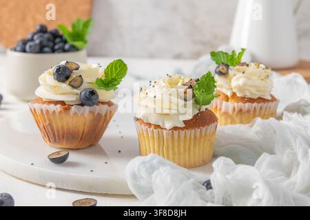 Three freshly baked cupcakes with white cream frosting, blueberries, and mint sit on a white marble board under bright natural light, creating an appe Stock Photo