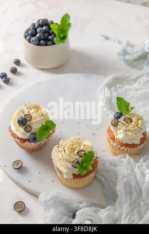 Three frosted cupcakes topped with fresh blueberries and mint rest on a round marble board, illuminated by soft natural light over a white textured su Stock Photo