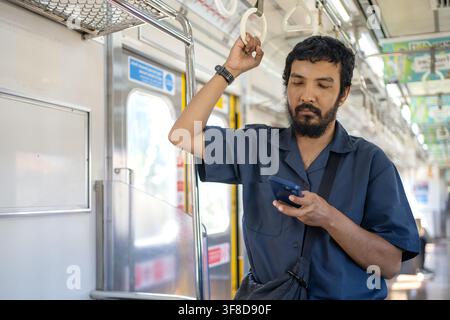 Male commuter standing inside a modern train while holding an overhead handrail and checking his smartphone during a daily public transportation journ Stock Photo