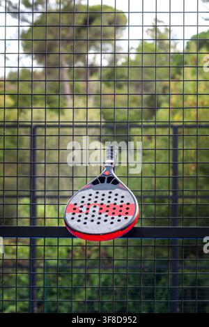 View of a padel racket resting against the black metal mesh fence of a padel court with lush green trees in the background in Cascais, Portugal. Stock Photo