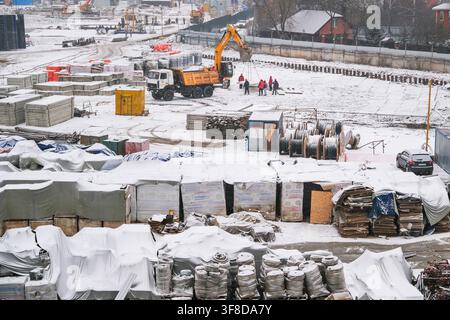 Moscow, Russia, 03.03.2025 Construction workers in red protective helmets supervising excavator and truck on snow covered building site during winter. Stock Photo