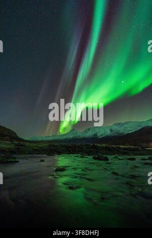 View of the vibrant northern lights illuminating the night sky above the snowy mountains and a calm river in Tromso, Troms, Norway. Stock Photo