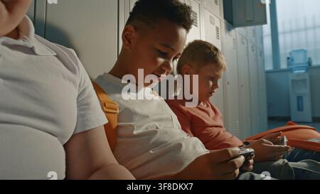 Children Enjoying Quiet Moment in School Hallway, Interacting Through Digital Devices while Sitting Beside Lockers. Concept of Modern Childhood, Education, Friendship, Diversity, Technology. Stock Photo