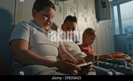 Generation Alpha Students Spending Free Time Together in School Hallway, Sharing Entertainment on Digital Devices while Relaxing Near Lockers. Friendship, Diversity, Education, Connected Childhood. Stock Photo