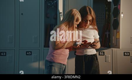Two Girls by Metal Lockers in Sunlit School Hallway, Both Engrossed in Own Smartphones. Concept of Generation Alpha, Modern Student Lifestyle Focusing on Screen Time, Social Media During School Break. Stock Photo