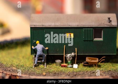 A close-up shows a miniature construction worker climbing a ladder beside a green corrugated cabin with tiny tools on grassy ground. Stock Photo