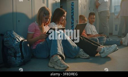Two School Girls Sitting on Floor Near Lockers, Working Together on Laptop and Digital Tablet During Break. Boys Sits Nearby, Focused on Smartphone. Digital Literacy, Modern Education Technology. Stock Photo