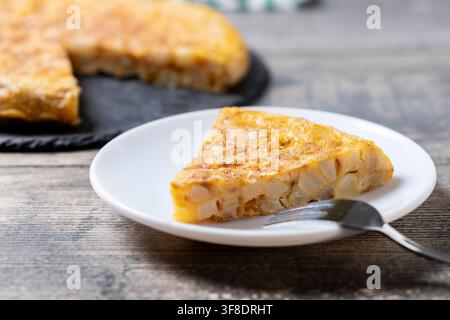 Slice of traditional Spanish potato omelette made with eggs and potatoes, served on a white plate with a fork on a rustic wooden table. Stock Photo