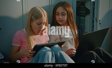 Two Focused Girls Using Personal Digital Devices for Collaborative Learning and Group Assignment in School Corridor Lined with Lockers. Educational Platforms, Technology Companies, STEM Education. Stock Photo