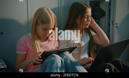 Two Focused Girls Using Personal Digital Devices for Collaborative Learning and Group Assignment in School Corridor Lined with Lockers. Educational Platforms, Technology Companies, STEM Education. Stock Photo