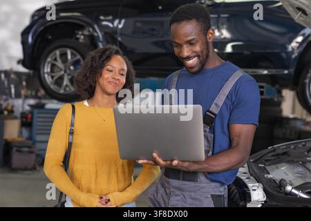 Professional mechanic in a workshop using a laptop for modern vehicle diagnostics, showing the woman client her car engine data. Stock Photo