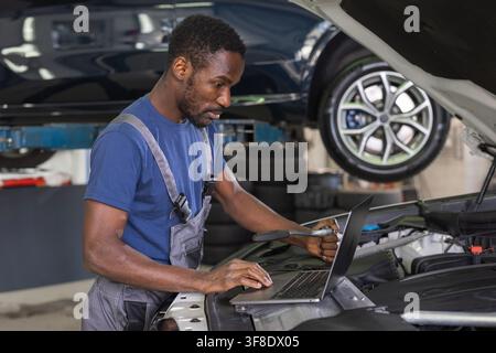 African American car mechanic in a modern repair shop performing vehicle maintenance with the hood open, using a laptop for diagnostics. Stock Photo