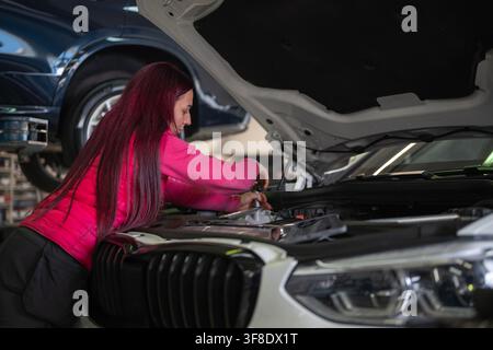 Focused female mechanic servicing a car engine under the hood, using a ratchet, hand tool for repairs. Women in skilled trades concept. Stock Photo