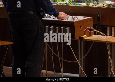 An adult man stands beside a wooden control panel with switches and cables in an indoor model railway setting. Stock Photo