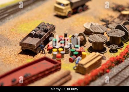 A close-up view of a miniature industrial diorama shows a flatbed trailer loaded with barrels beside scattered drums, cable spools, and tiny worker fi Stock Photo