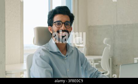 Successful Arabian Indian business man working on laptop looking at camera indoor office entrepreneur typing project startup smiling muslim Stock Photo