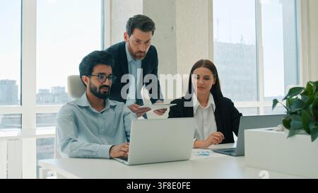 Male CEO business leader helping office coworkers with laptop project indoors team meeting boss man guide multiethnic colleagues businesswoman Arabian Stock Photo