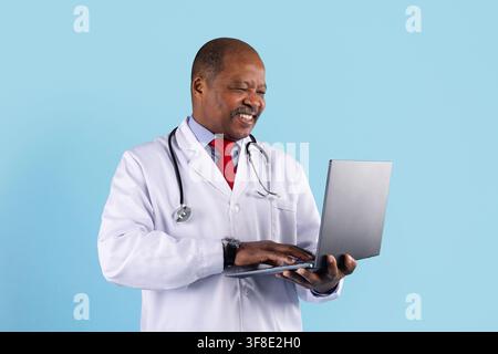 Smiling mature African American doctor using laptop on blue studio background Stock Photo