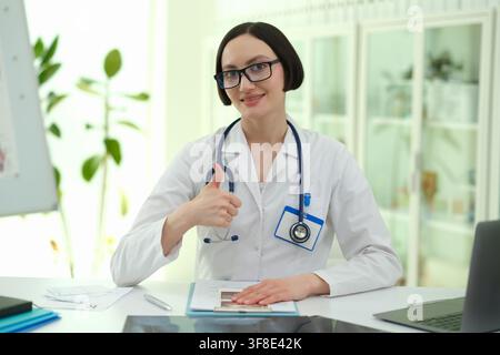 Smiling female doctor gives thumbs up as sits in clinic workspace Stock Photo