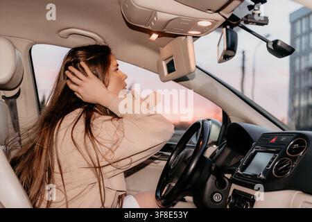 Woman car mirror young driver in trench coat adjusts hair looking in visor mirror during a golden hour commute Stock Photo