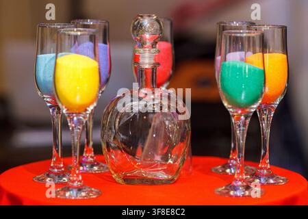 A glass decanter and multiple champagne glasses filled with colorful sand stand on a red table for a sand ceremony. Stock Photo