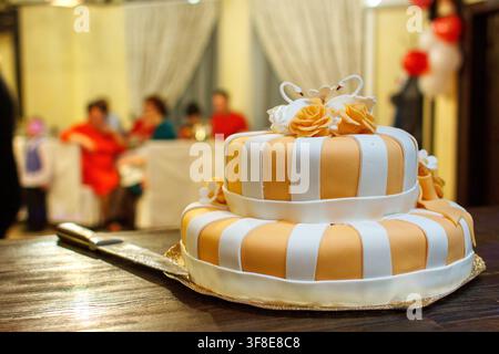A two-tiered wedding cake with orange and white stripes, topped with swans and roses, sits on a table. Stock Photo