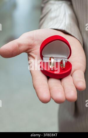 A man holds an open red velvet box containing two golden wedding rings in the palm of his hand. Stock Photo