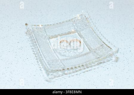 Two wedding rings rest in the center of an ornate square crystal dish on a light surface. Stock Photo