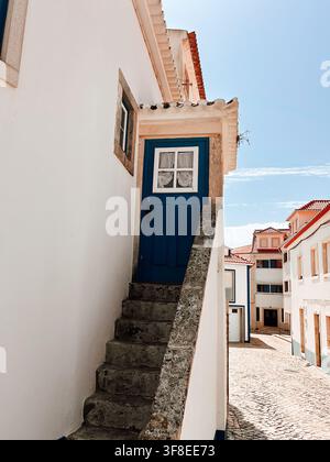 Traditional blue wooden door reached by a narrow exterior stone staircase on a whitewashed house, with tiled roofs and cobblestone street in the backg Stock Photo