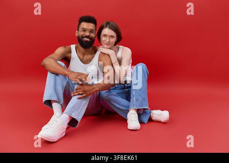 A joyful couple sits on a vibrant red background, expressing love and connection in their outfits. Stock Photo