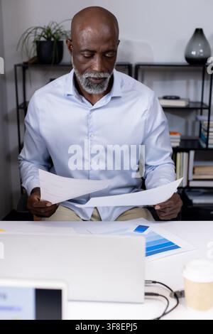 Senior man in blue shirt reading papers and color chart at office desk with laptop. Workspace, shelf, plant, volumes, documents, bargraph, minimalist Stock Photo