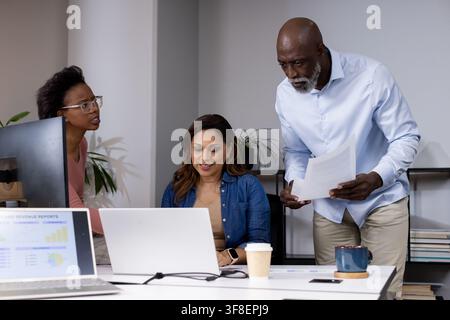 Three colleagues leaning over desk, reviewing white laptop and printed reports at modern office Stock Photo