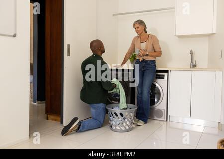 Diverse couple kneeling and standing, loading washer and dryer in laundry room with basket Stock Photo
