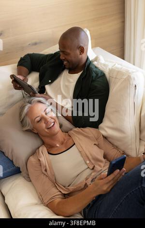 Diverse couple reclining on light-colored sofa in living room, using tablet and smartphone Stock Photo