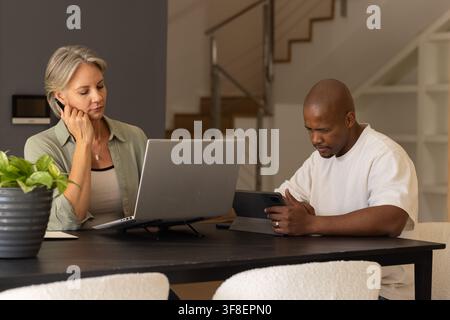 Diverse couple sitting at dining table at home using laptop and tablet with planter Stock Photo