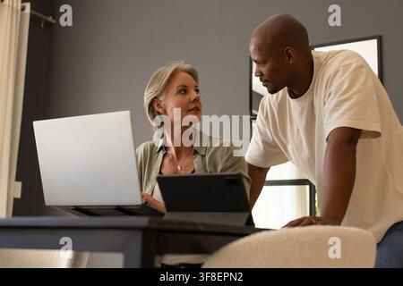 Diverse couple working and conversing at home dining table with silver laptop on stand and tablet Stock Photo