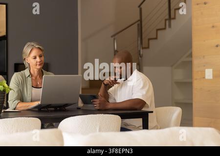 Couple sitting at dining table at home using laptop on riser and tablet, woman olive overshirt Stock Photo