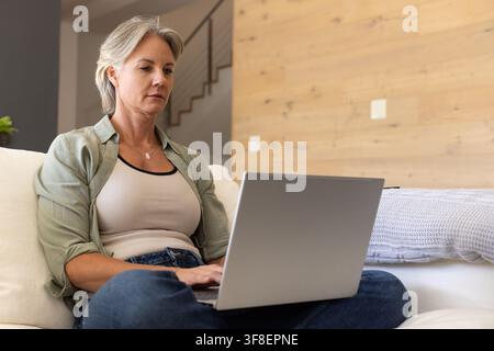 Senior female wearing light-green shirt sitting on sofa in living room typing on laptop, copy space Stock Photo